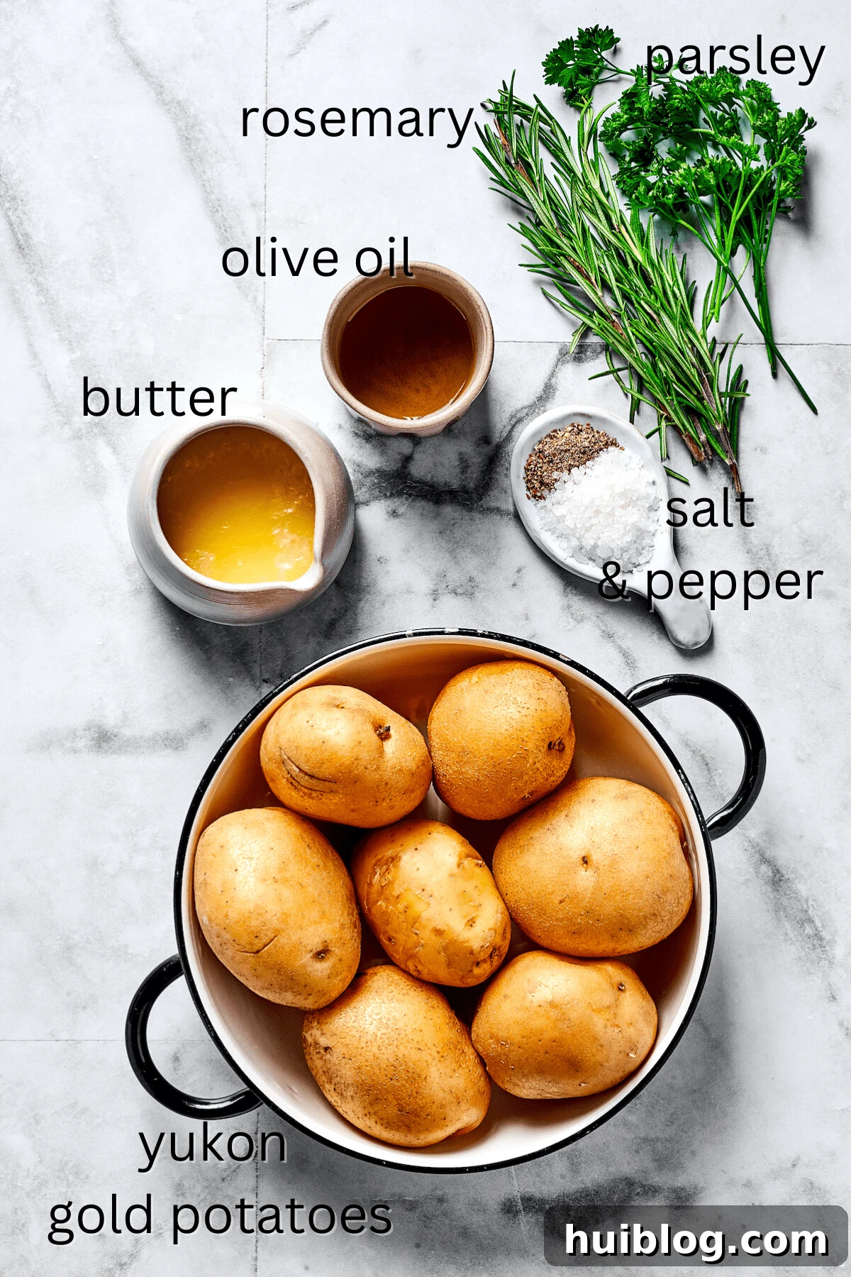 A flat lay of ingredients for roasted potatoes: fresh herbs, olive oil, melted butter, salt, pepper, and cut potatoes.