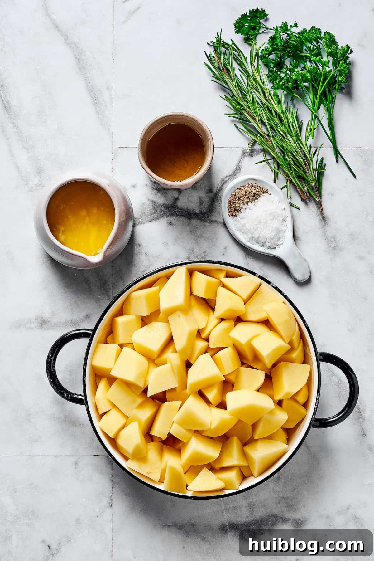 A flat lay of fresh ingredients including olive oil, fresh rosemary, fresh parsley, melted butter, salt and pepper, and cut-up raw potatoes.