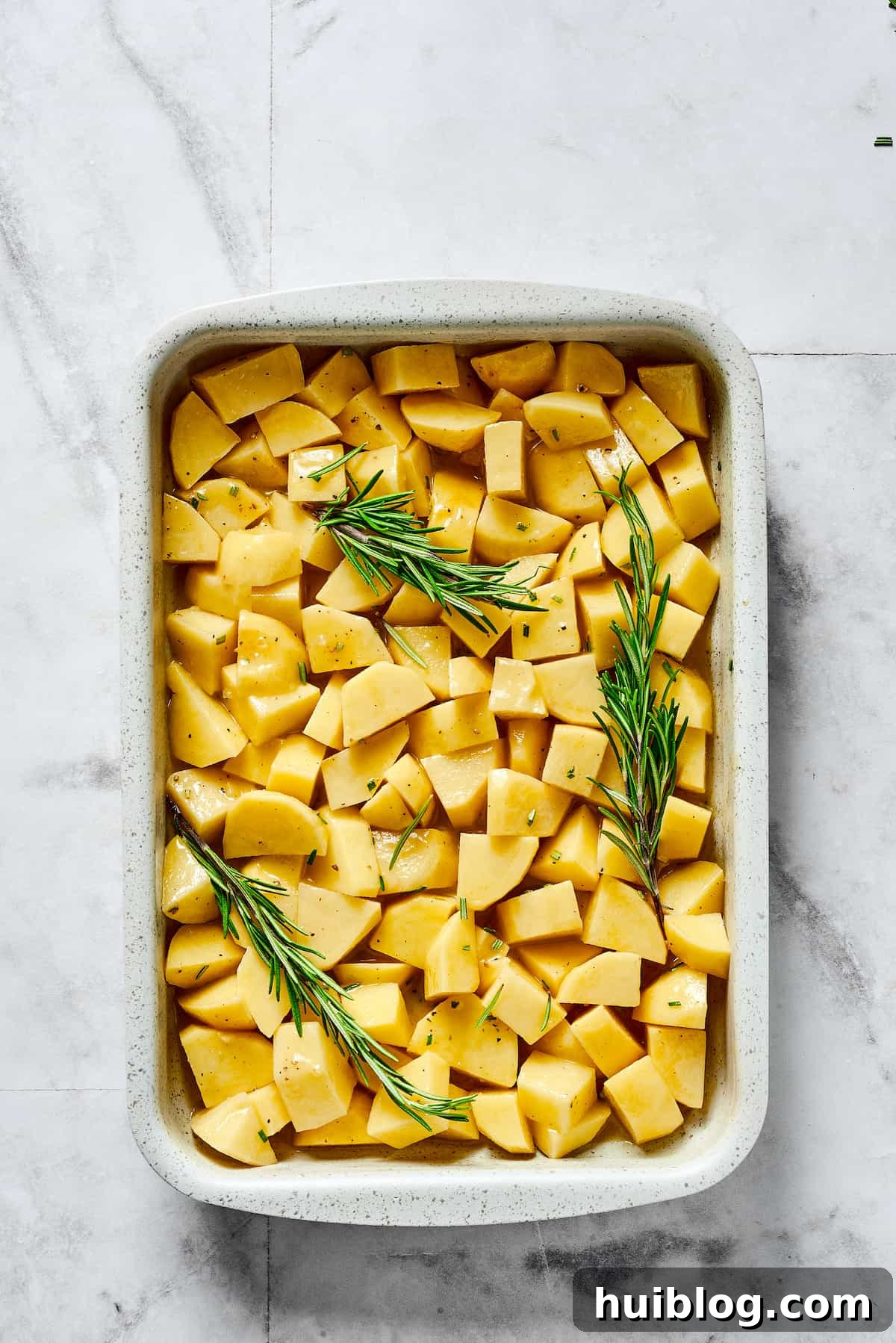Seasoned raw potatoes and rosemary sprigs spread evenly in a baking dish, ready to be roasted.