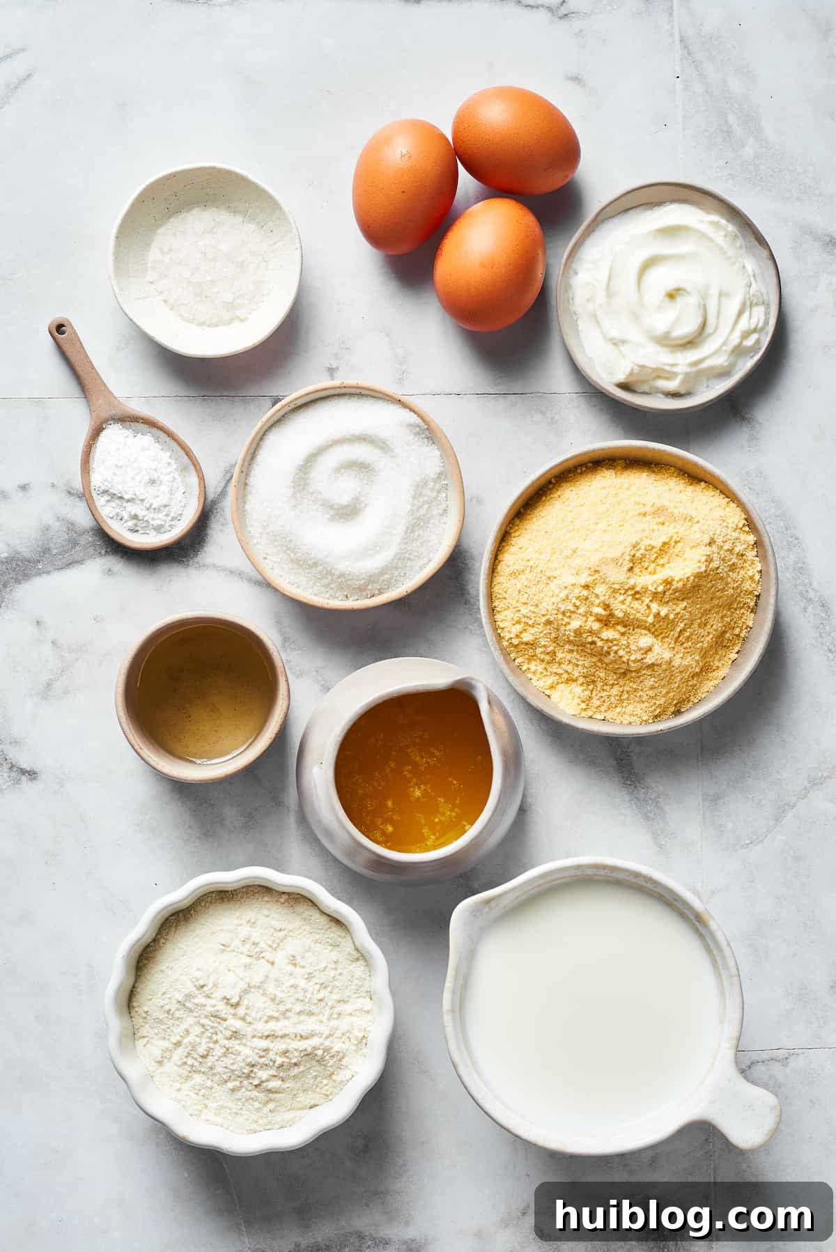 An overhead shot showcasing all the essential cornbread ingredients neatly arranged: salt, eggs, sour cream, baking powder, sugar, cornmeal, oil, melted butter, flour, and milk.