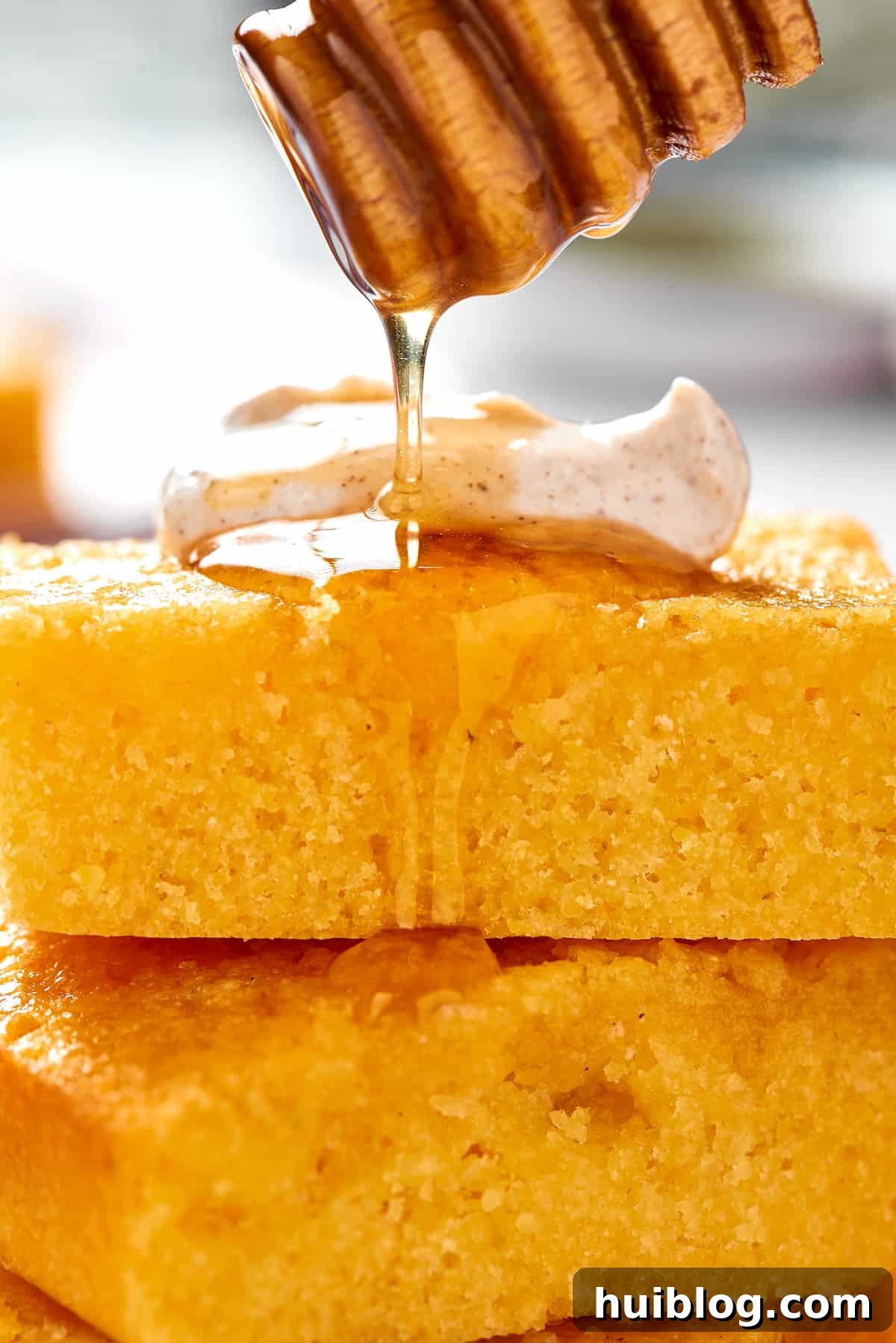 A honey dipper gracefully drizzles golden honey over squares of freshly baked cornbread, some topped with fluffy cinnamon cream, highlighting its sweet appeal.