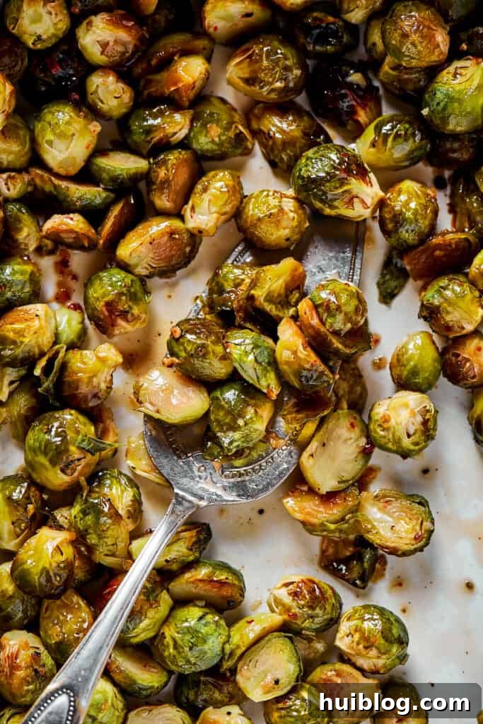 A close-up of roasted Brussels sprouts being gently tossed with the rich, shiny maple-pomegranate glaze on a baking sheet, ensuring every sprout is coated.