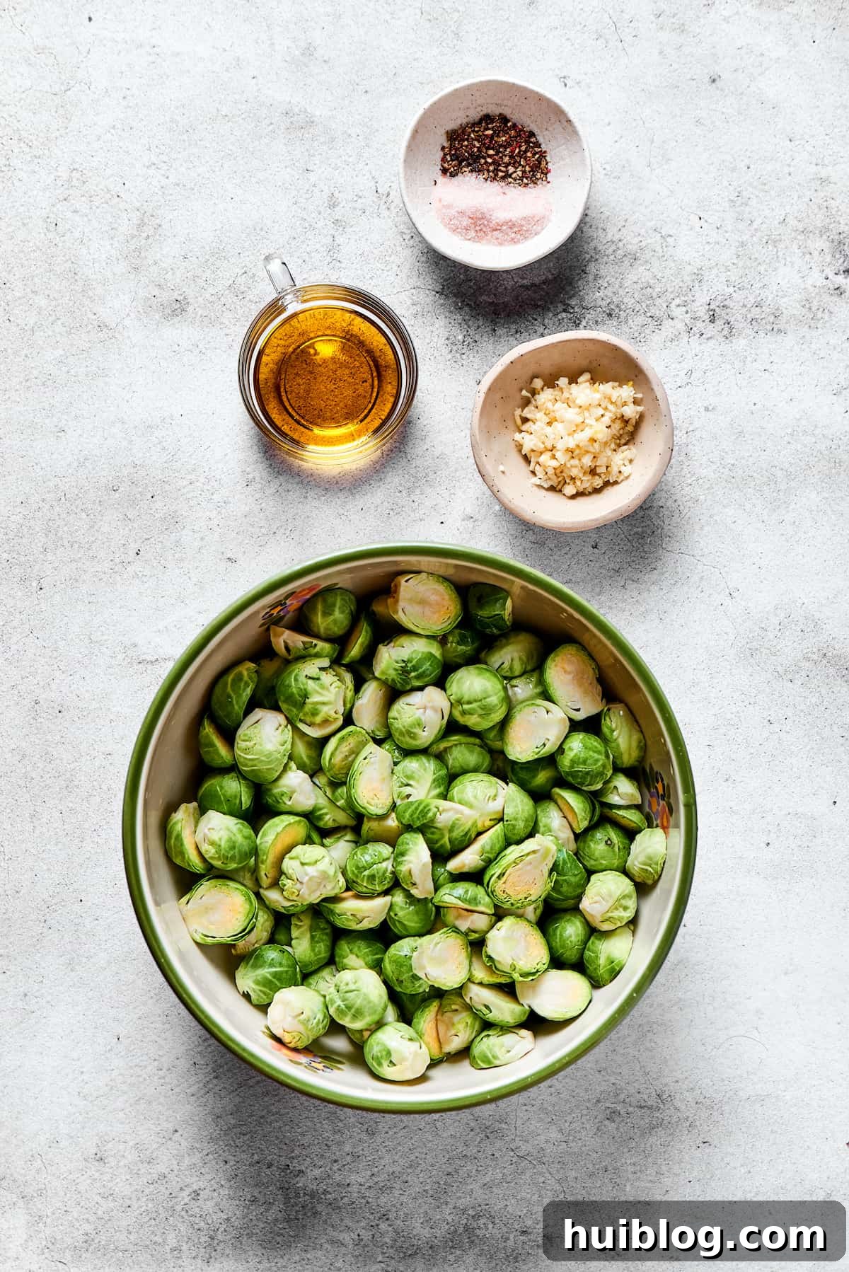Top down view of ingredients: Fresh Brussels sprouts, olive oil, minced garlic, and shakers of salt and pepper on a rustic wooden surface.