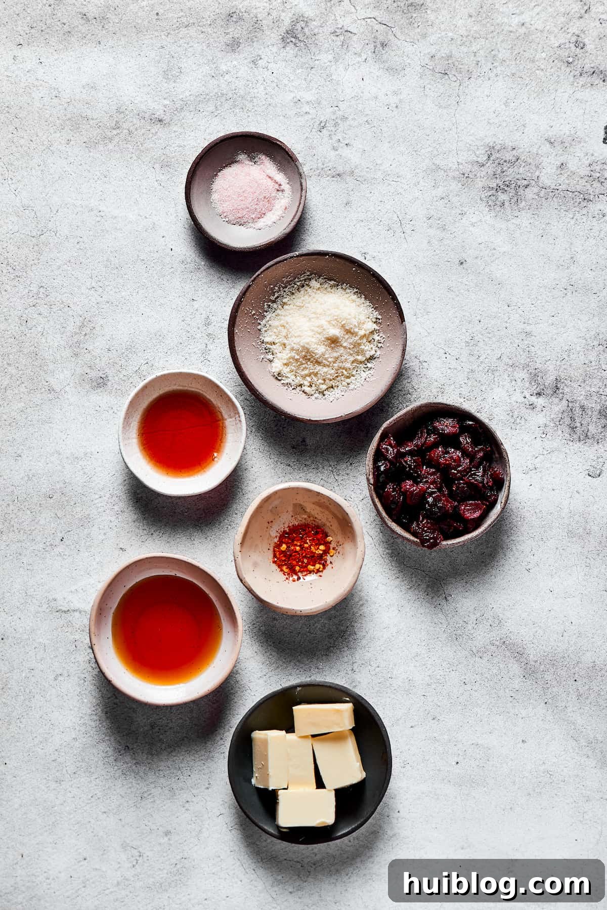 Top down view of glaze ingredients: A knob of butter, a bowl of red chili flakes, dried cranberries, grated Parmesan cheese, a bottle of maple syrup, and pomegranate vinegar.