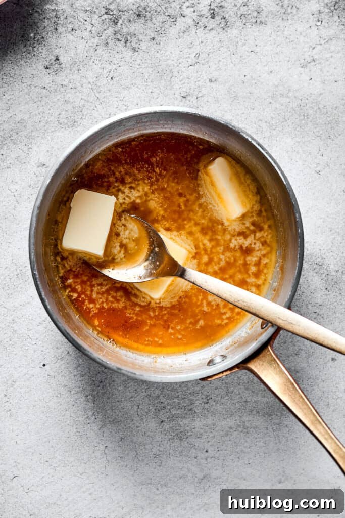A close-up shot of butter being stirred into the simmering maple-pomegranate glaze in a small saucepan, creating a rich, glossy texture.