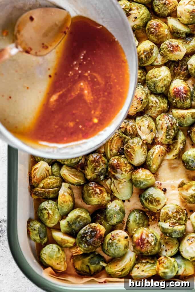Pouring the glossy maple-pomegranate glaze over a baking sheet full of freshly roasted Brussels sprouts, ready to be tossed.