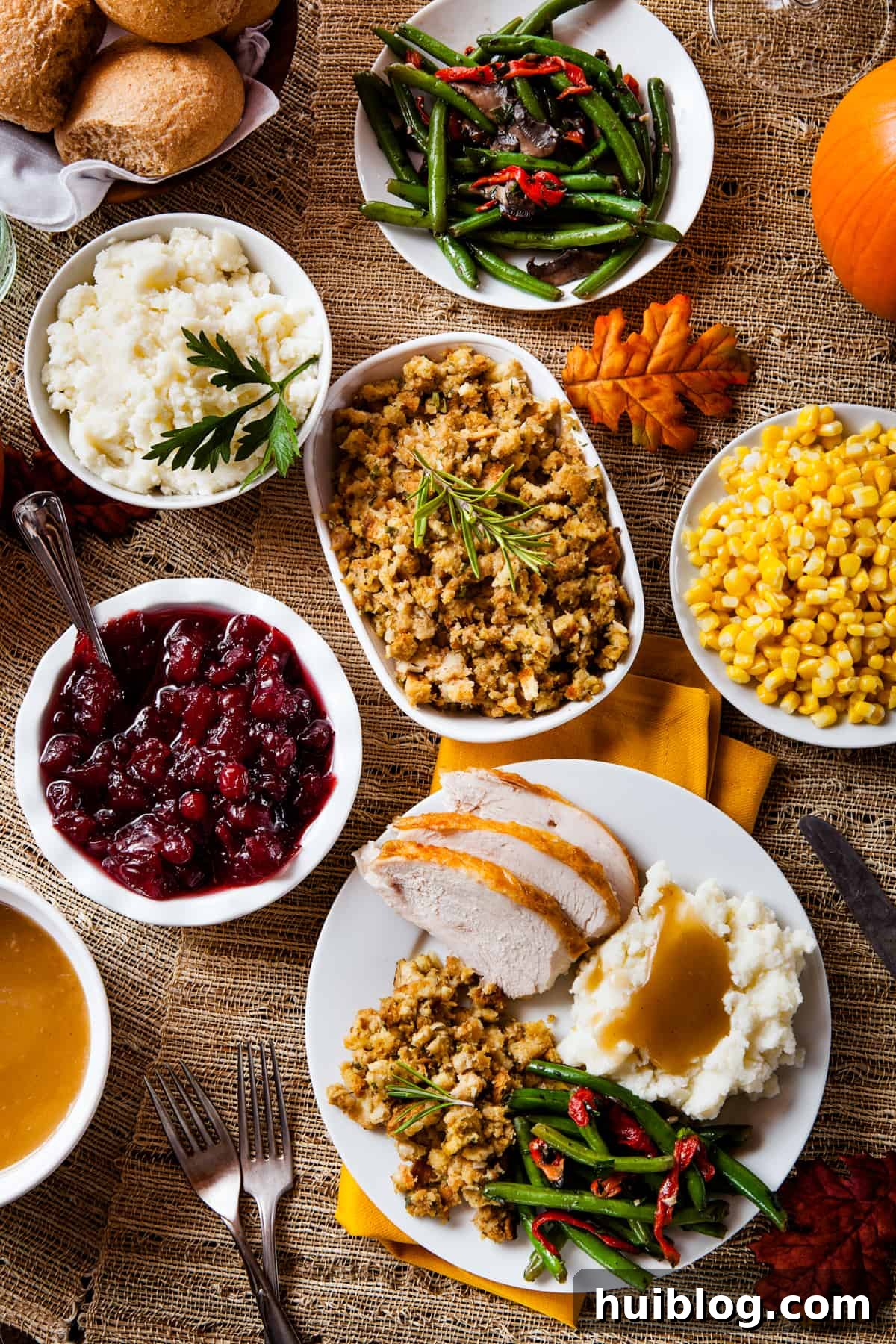 Classic Roast Turkey with Stuffing, fresh herbs, and a pretty pumpkin garnish is easy to make from scratch. The perfect Turkey Day feast! An inviting overhead shot displaying a variety of Thanksgiving side dishes arranged on a table. Featured are a gravy boat, cranberry sauce in a white bowl, creamy mashed potatoes, savory stuffing, crisp green beans, sweet corn, and a plate with succulent sliced turkey alongside mashed potatoes, green beans, and corn.