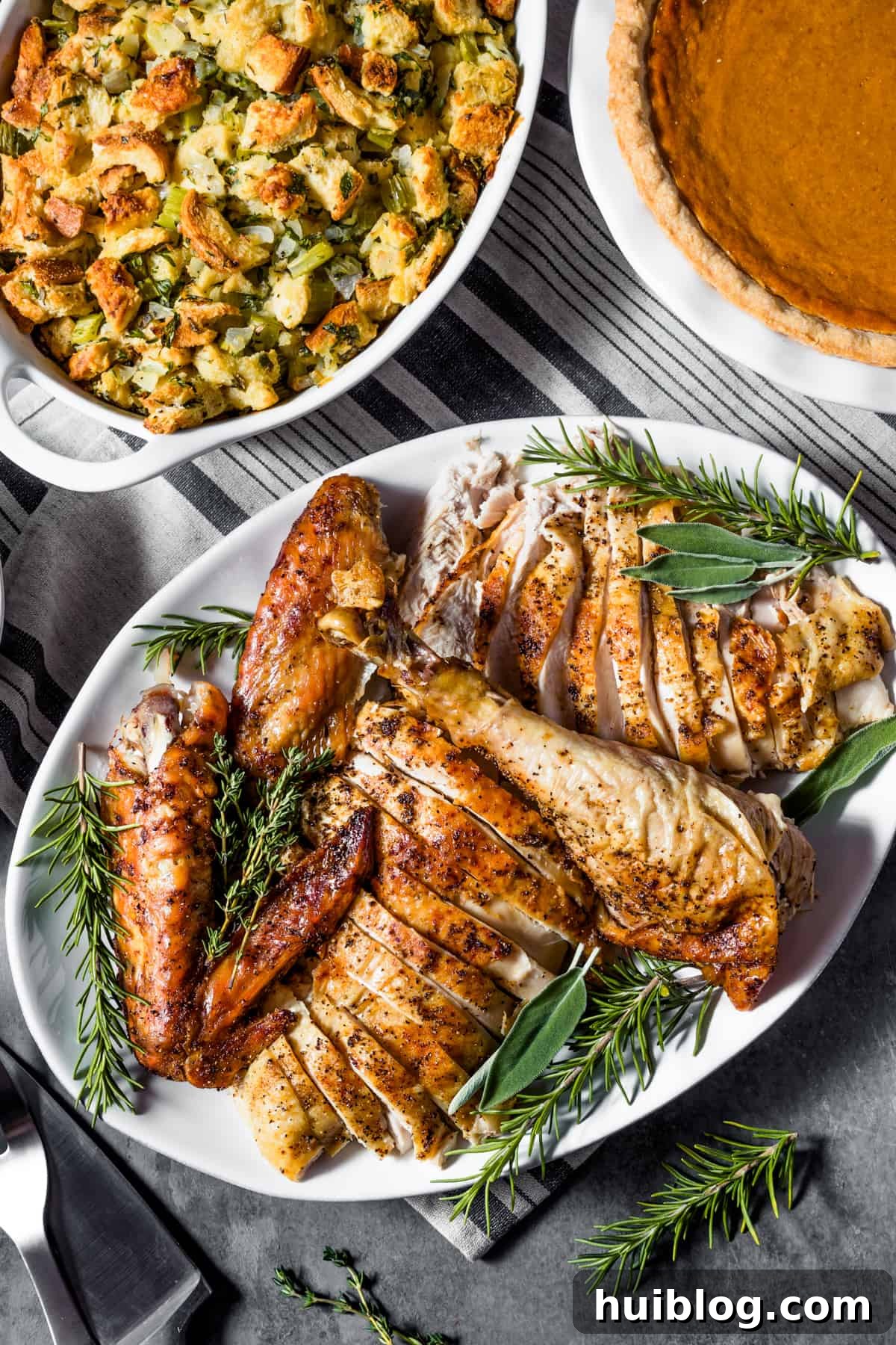 Classic Roast Turkey with Stuffing, fresh herbs, and a pretty pumpkin garnish is easy to make from scratch. The perfect Turkey Day feast! An inviting overhead shot showcasing a beautifully carved roast turkey served on a white oval platter, elegantly garnished with fresh herbs. Just above the turkey, a bowl of savory stuffing and a slice of delicious pumpkin pie complete the festive holiday spread.