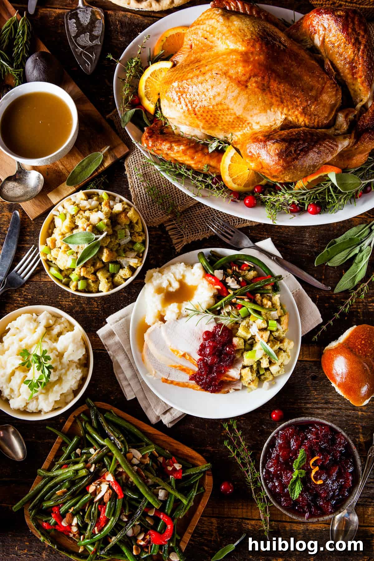 Classic Roast Turkey with Stuffing, fresh herbs, and a pretty pumpkin garnish is easy to make from scratch. The perfect Turkey Day feast! An appetizing overhead shot featuring a whole roast turkey at the top of the frame, surrounded by an array of classic holiday side dishes: a bowl of creamy mashed potatoes, savory stuffing, fresh green beans, and a vibrant cranberry sauce.