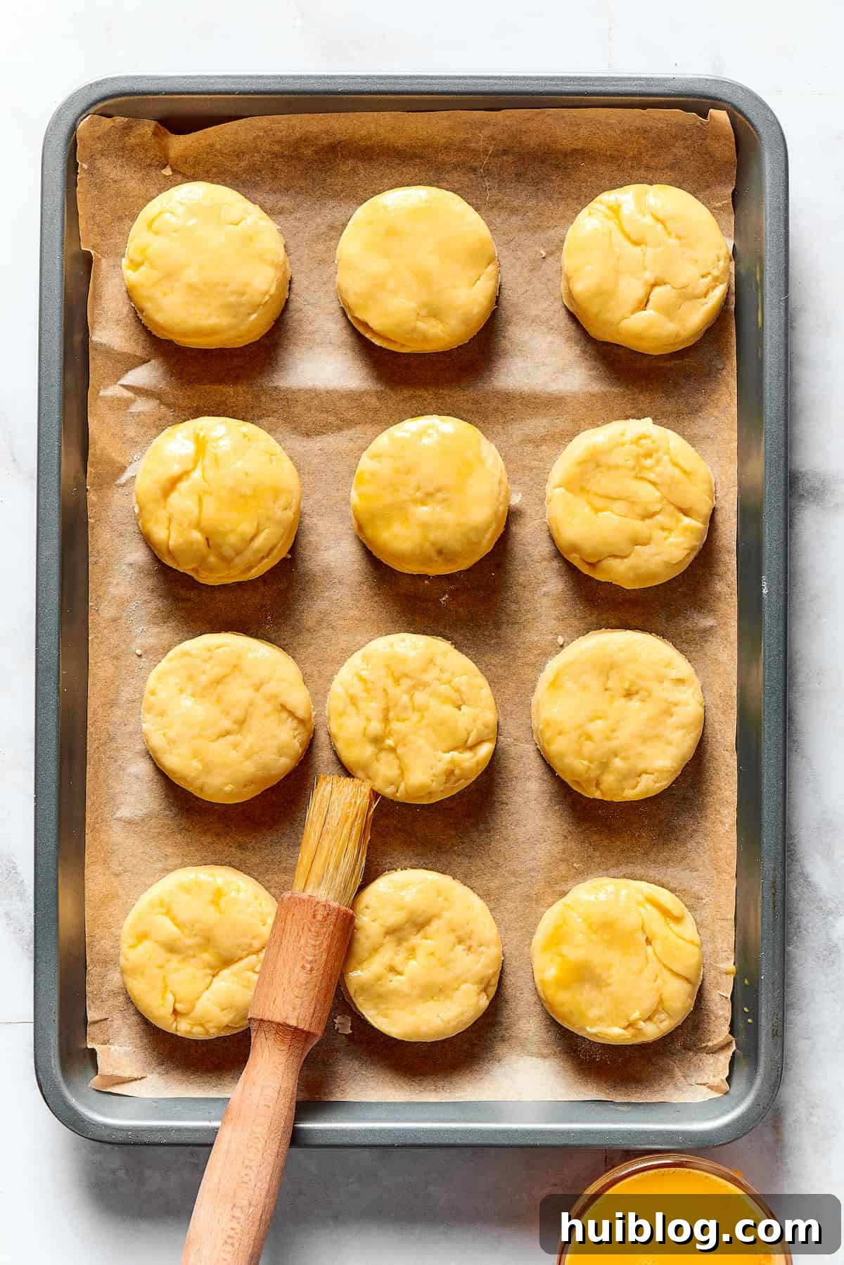 A dozen unbaked biscuit rounds neatly arranged on a baking sheet, being brushed with beaten egg for a golden glaze before baking.