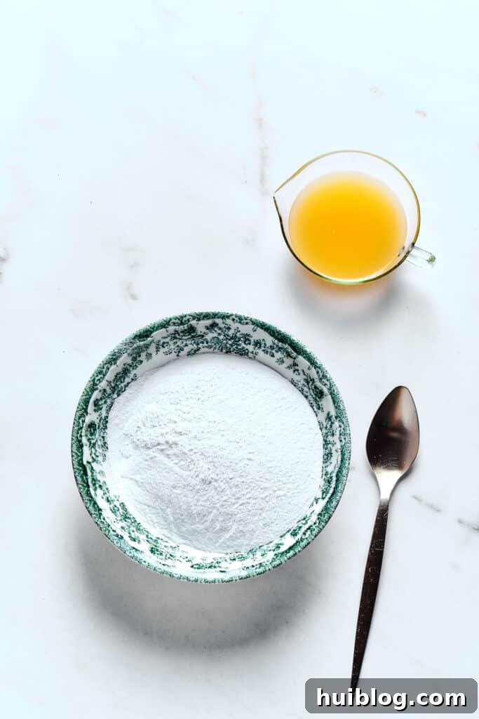 A bowl of powdered sugar beside a glass of fresh orange juice and a spoon, ready for making the glaze.