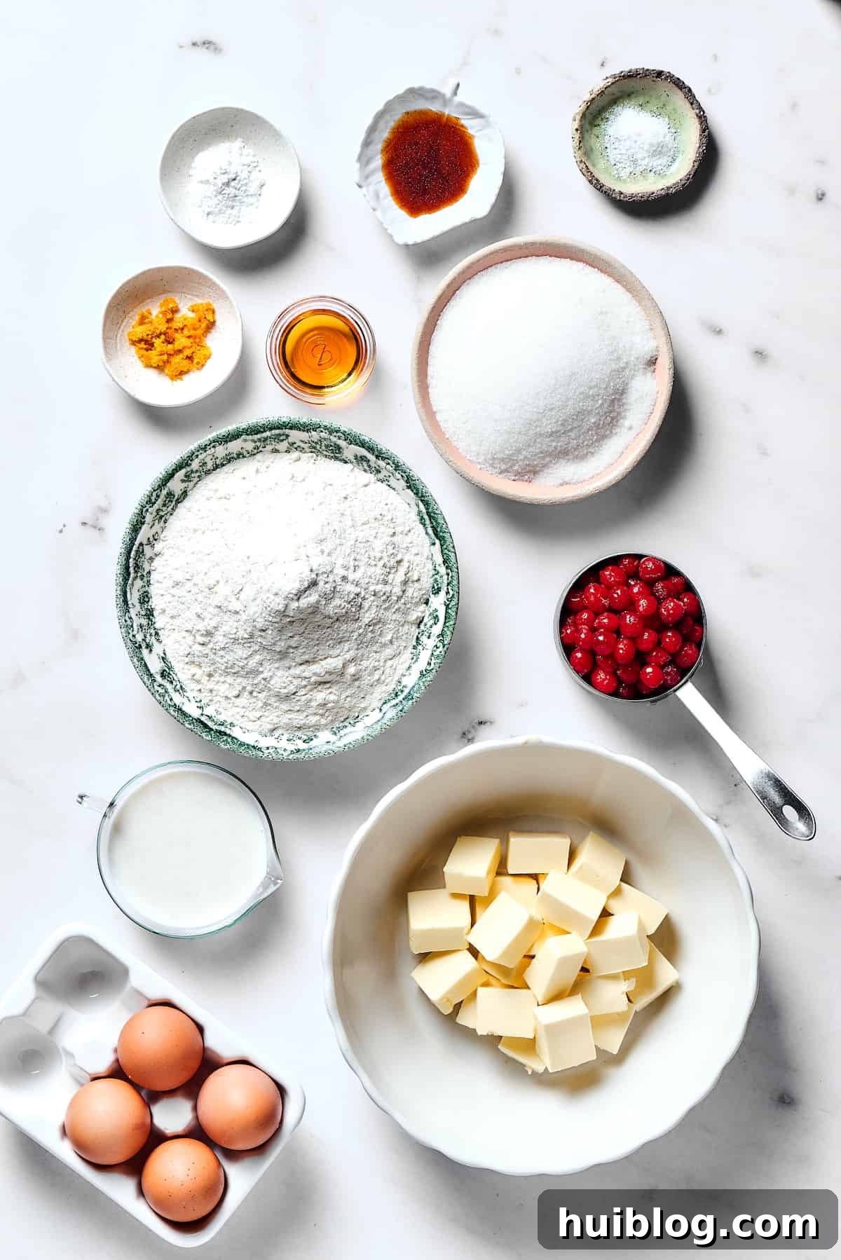 A collage of fresh ingredients laid out on a table: baking powder, vanilla bean paste, salt, orange zest, orange liqueur, granulated sugar, all-purpose flour, fresh cranberries, buttermilk, unsalted butter, and large eggs.