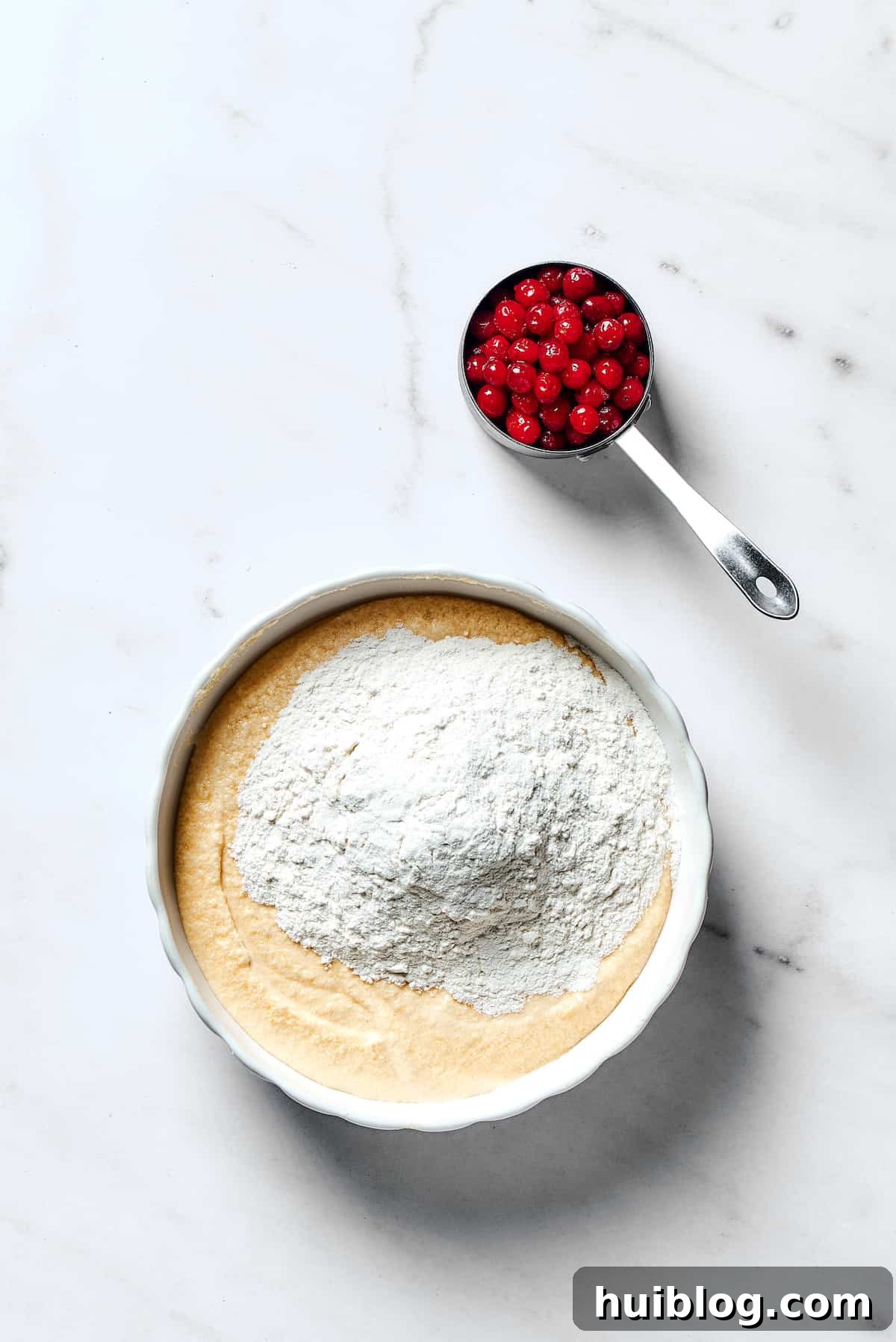 Dry ingredients being incorporated into the wet batter in a mixing bowl, with a measuring cup of fresh cranberries ready to be folded in.