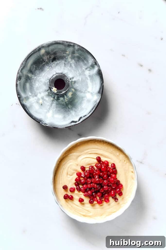 A buttered and floured Bundt pan sits next to a bowl of cake batter, with fresh cranberries sprinkled on top of the batter.