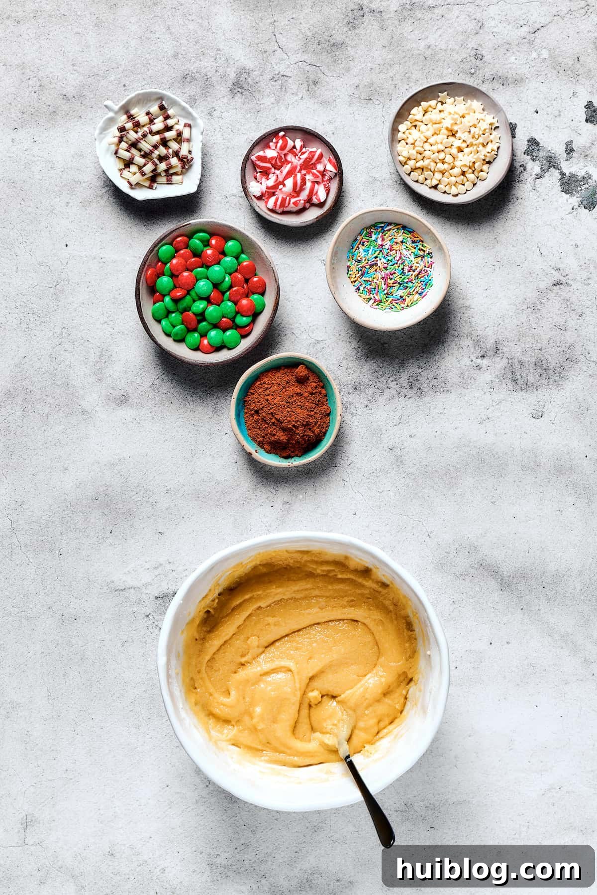 A large bowl of creamy cookie dough sits invitingly on a wooden table, flanked by small bowls brimming with festive red and green M&Ms and white chocolate chips, ready for cookie decoration.
