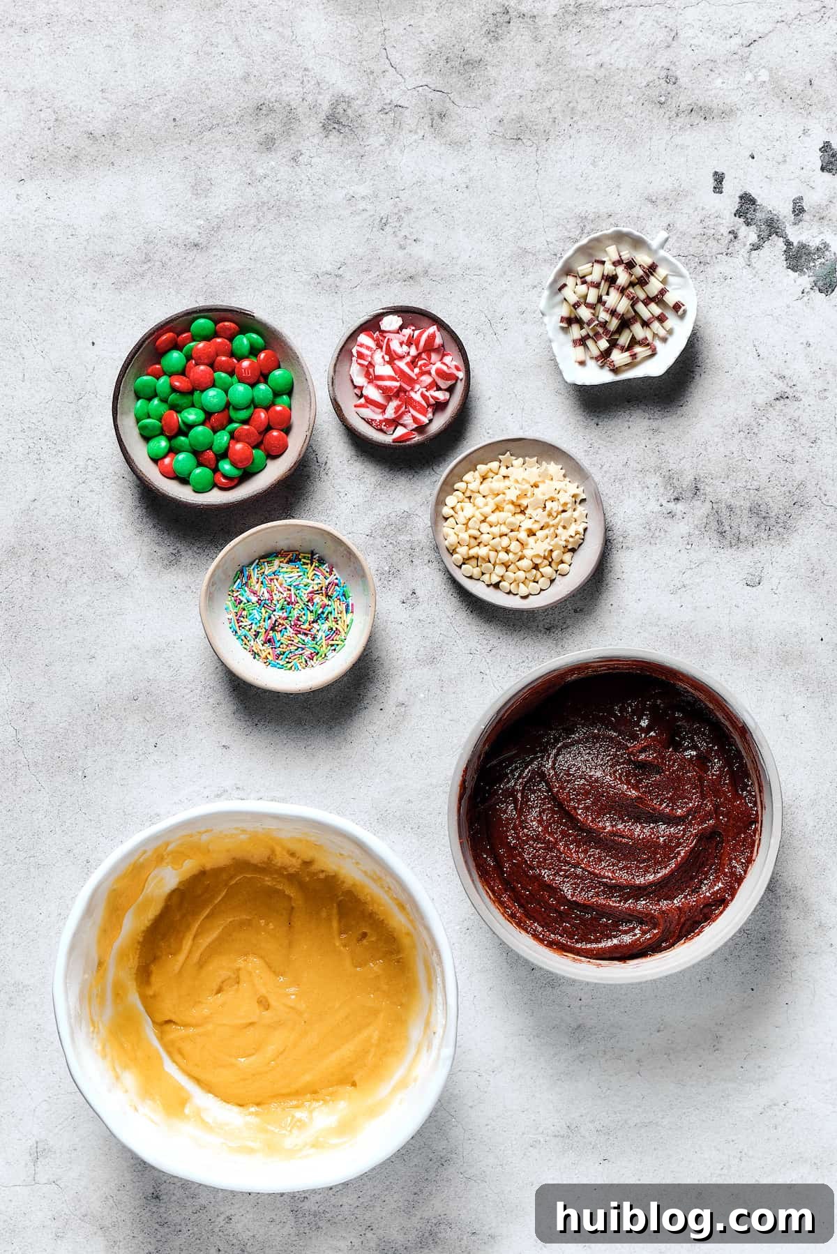 Two bowls of perfectly prepared cookie dough, one rich chocolate and one classic vanilla, sit side-by-side on a wooden surface, surrounded by small dishes of festive decorations like M&Ms and sprinkles.