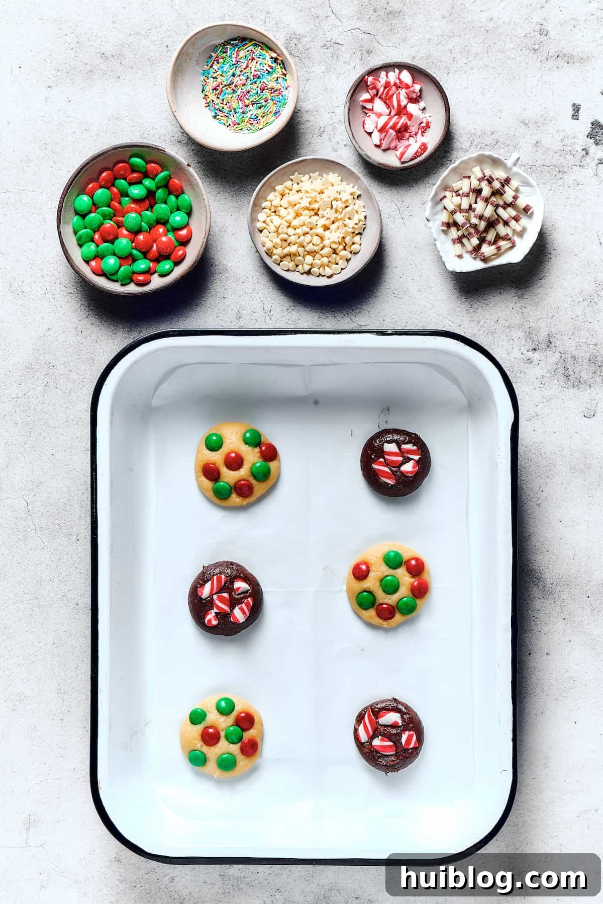 An assortment of unbaked chocolate and vanilla cookies, generously topped with colorful M&M's, arranged neatly on a parchment-lined baking sheet, ready for the oven.