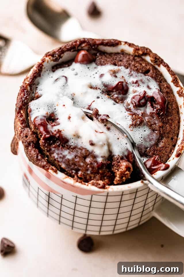 A close-up shot of a spoon scooping into a fluffy chocolate mug cake, generously topped with whipped cream and chocolate shavings.