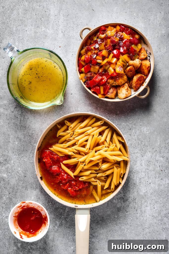 Penne pasta, crushed tomatoes, and chicken broth simmering together in a skillet, alongside a separate dish of cooked chicken and vegetables.