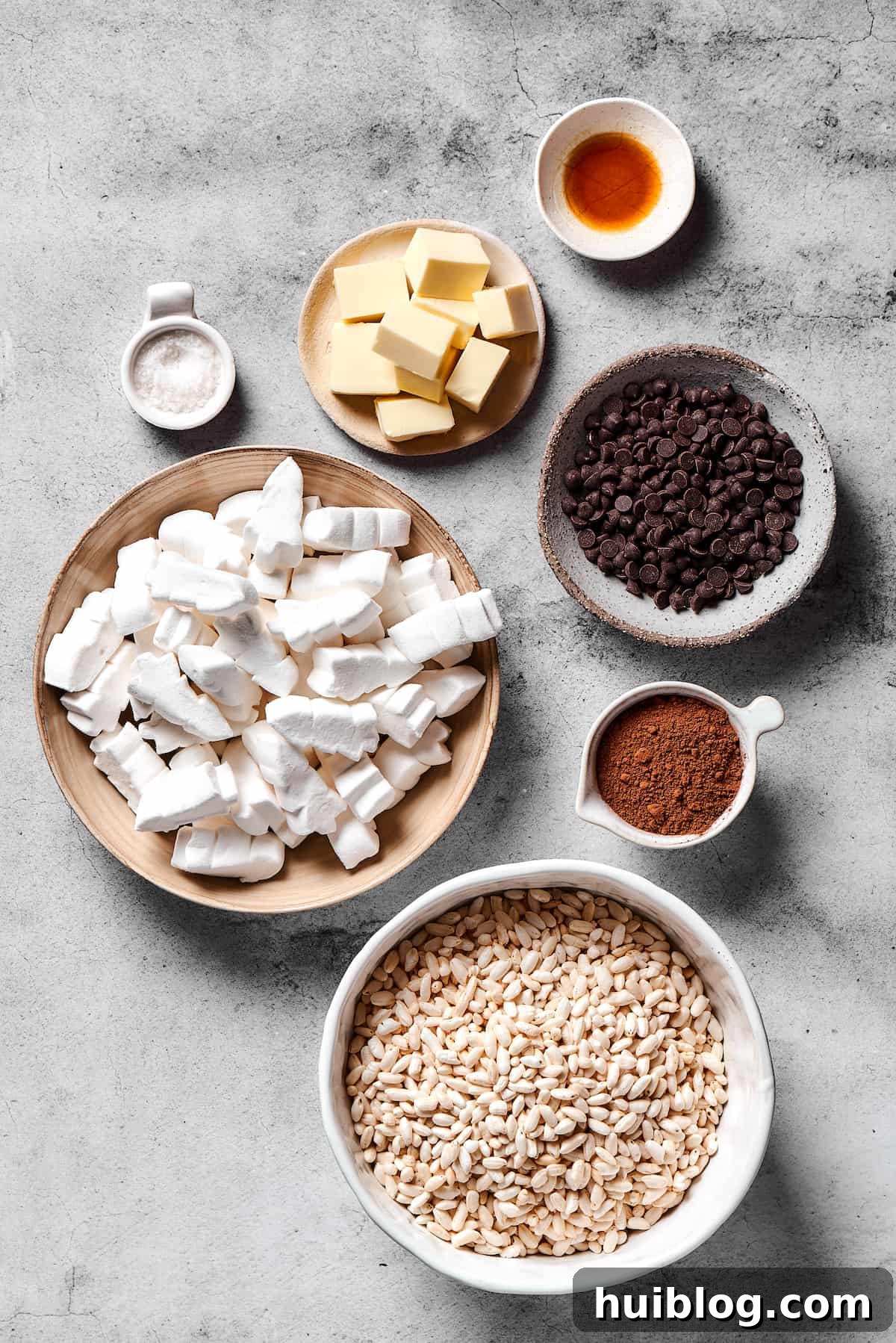 All the essential ingredients for making delightful chocolate rice krispie treats, laid out for preparation. An array of ingredients for Chocolate Rice Krispie Treats: vanilla extract, chocolate chips, cocoa powder, crispy rice cereal, marshmallows, butter, and flaky sea salt.