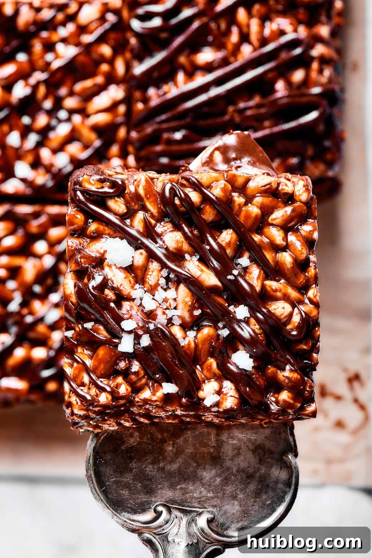 A tempting chocolate rice krispie treat being served, showcasing its perfect square shape and appealing texture. A silver dessert server gently lifting a square of chocolate cereal bar, highlighting its gooey texture and ready-to-eat appeal.