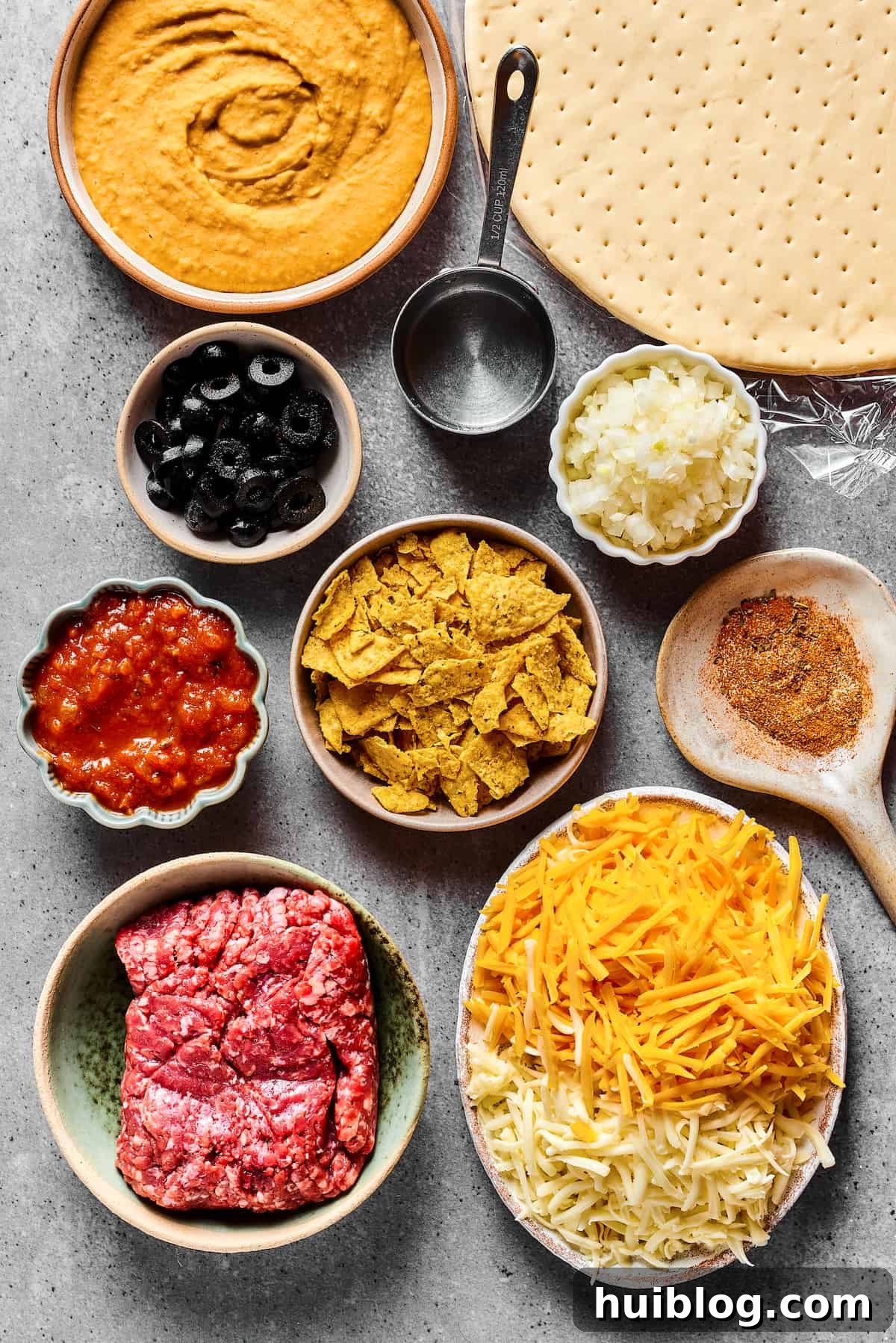 All the Fresh Ingredients for Homemade Taco Pizza A visual representation of all ingredients laid out, including refried beans, pizza crust, black olives, water, onion, salsa, crushed tortilla chips, taco seasoning, ground beef, and cheese, ready for assembly.