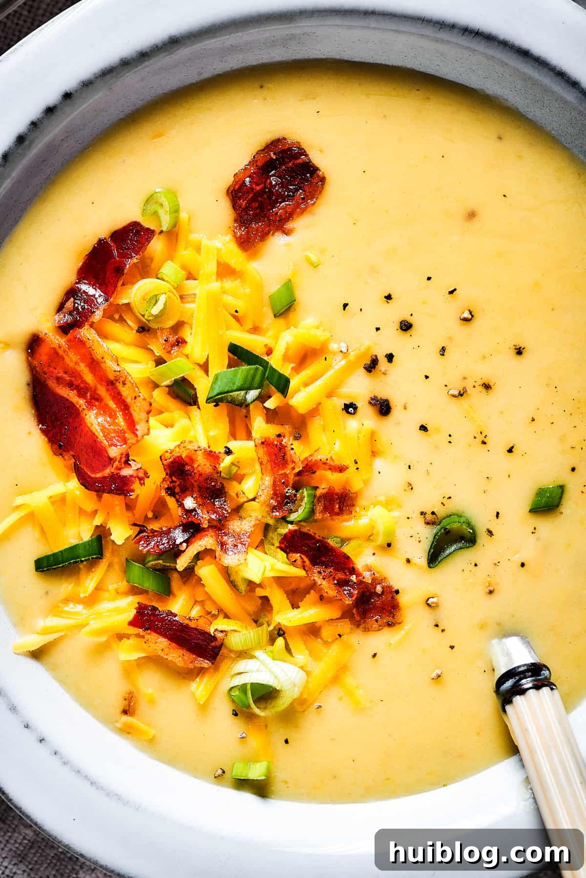 A close-up view of a bowl of creamy baked potato soup, adorned with a vibrant medley of crispy bacon, fresh green onions, and melted shredded cheese.
