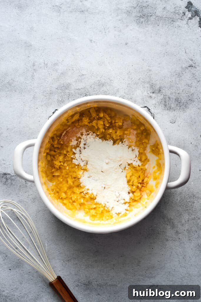 Flour being sprinkled into a soup pot, mixing with the cooked onions, butter, and garlic to form a smooth roux.