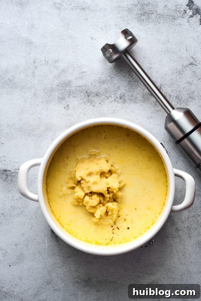 A large soup pot containing mashed potatoes immersed in a creamy soup base, with an immersion blender resting nearby, ready to create a smooth texture.