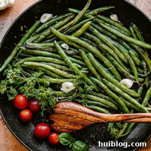 green beans, tomatoes, and garlic cloves in a black skillet with a wooden spoon inside