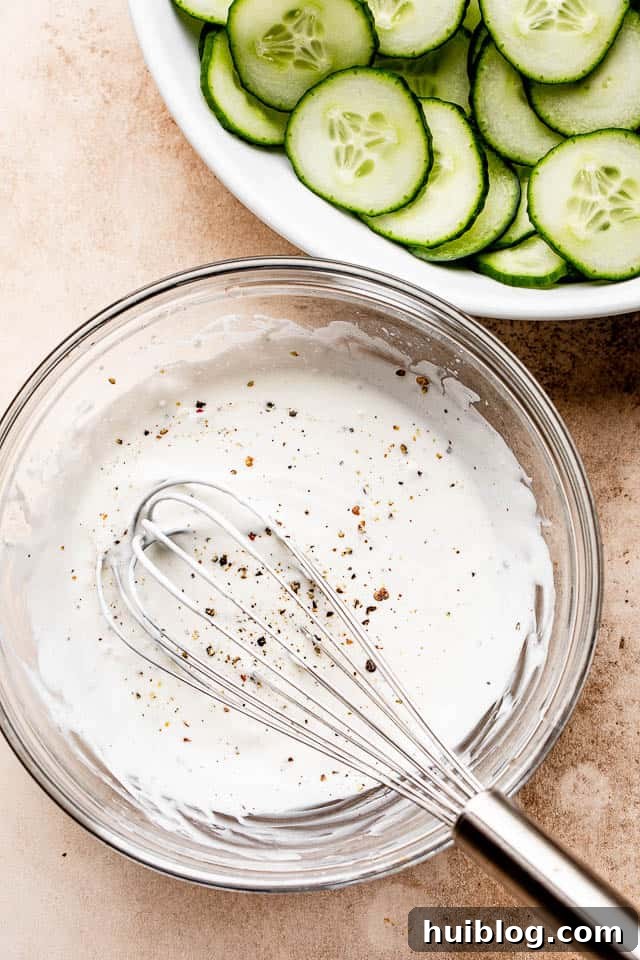 A smooth, tangy sour cream dressing expertly whisked in a clear glass bowl, with a whisk still inside, ready to be poured over fresh cucumbers.