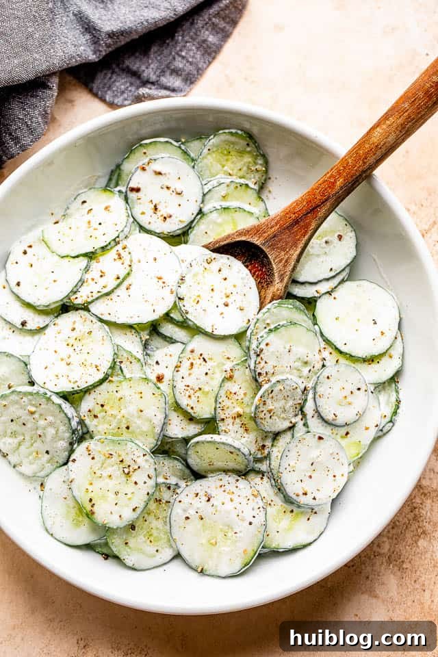 A wooden spoon gently mixing fresh cucumber slices in a white salad bowl, thoroughly coated with a creamy sour cream dressing.