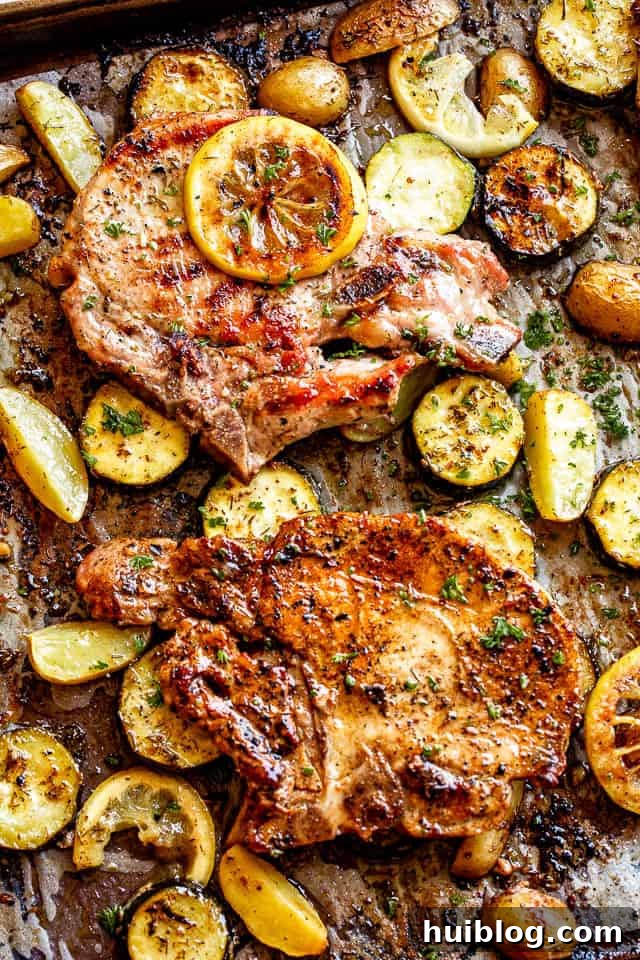 Golden brown baked pork chops resting on a bed of roasted squash and potatoes, garnished with fresh lemon slices, on a sheet pan