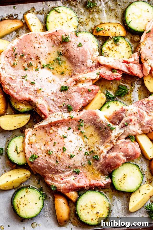 Raw, marinated bone-in pork chops laid out on a baking sheet over a single layer of sliced zucchini and quartered baby potatoes