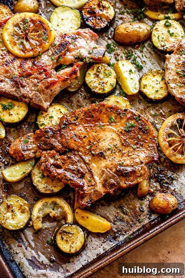A sheet pan with a complete meal of baked pork chops, roasted squash, and potatoes, garnished with fresh herbs and lemon slices