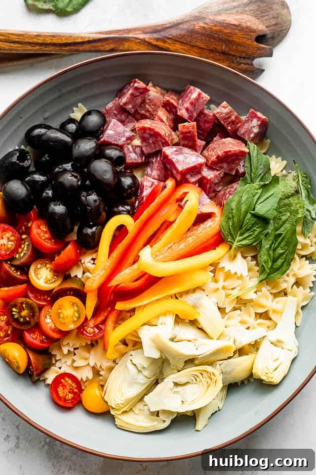 Italian Deli Pasta Salad 3 An overhead view of a wooden cutting board next to a salad bowl, showcasing arranged ingredients like pasta, olives, salami, cheese, artichokes, tomatoes, basil, and peppers.
