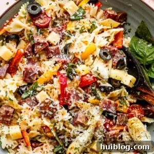 Top view of a wooden spoon in a bowl filled with pasta, cheese cubes, salami cubes, tomatoes, and olives.
