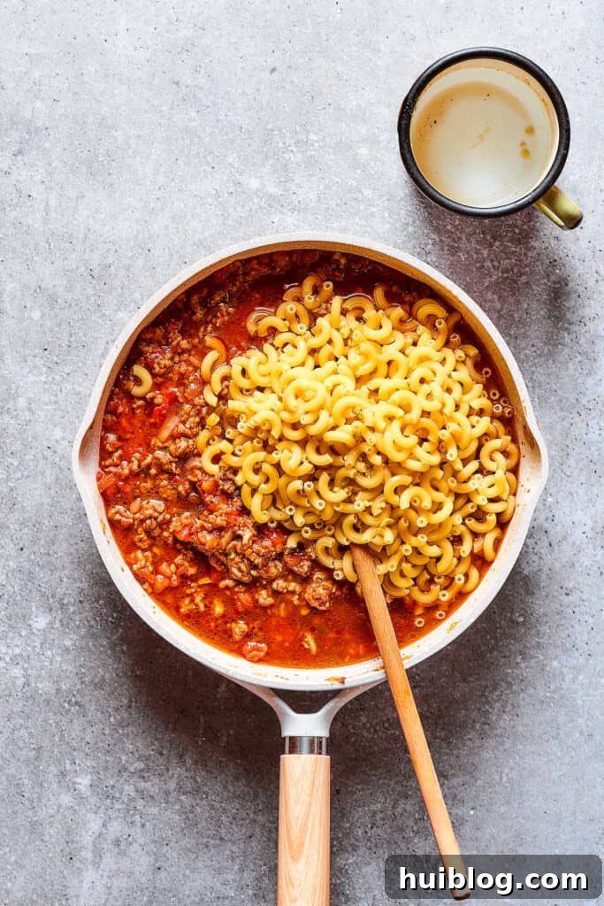 Incorporating dry macaroni pasta into the savory beef and tomato sauce mixture in the skillet.
