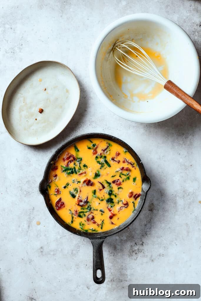 Pouring the whisked egg mixture over bacon, onion, spinach, and potatoes in a cast-iron pan, ready for cooking.