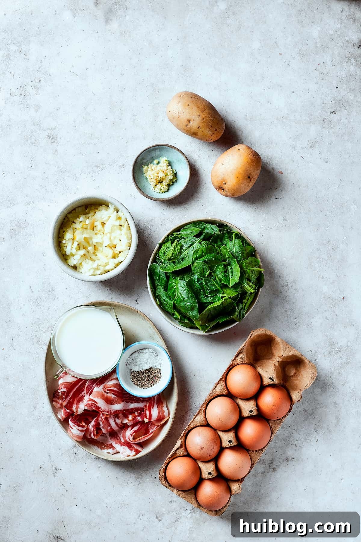 Individual ingredients laid out: Potatoes, fresh spinach, eggs, crispy bacon, salt and pepper, milk, diced onion, and minced garlic.