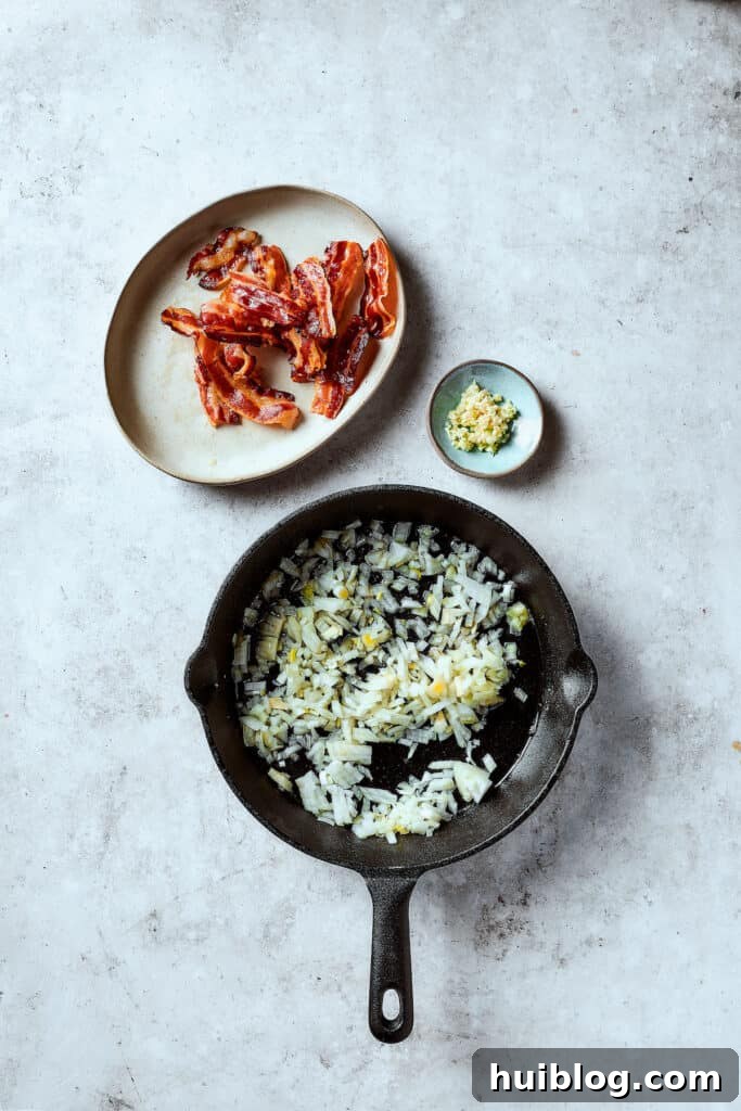 Frying diced onions in a skillet with bacon grease. A small dish of minced garlic and a plate of cooked bacon are nearby.