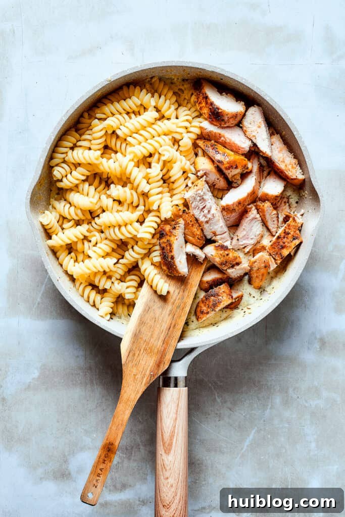 Combining chicken and pasta with the creamy sauce. Cooked chicken strips and al dente pasta being added to the creamy sauce in a skillet, ready to be combined.