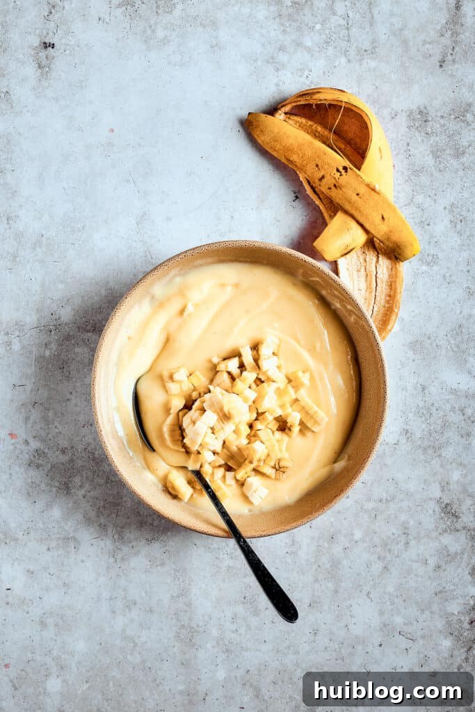 Incorporating fresh bananas into the pudding for a true banana pudding experience. Small pieces of chopped banana being stirred into the cooling pudding, ensuring every spoonful will have a burst of fresh fruit flavor.