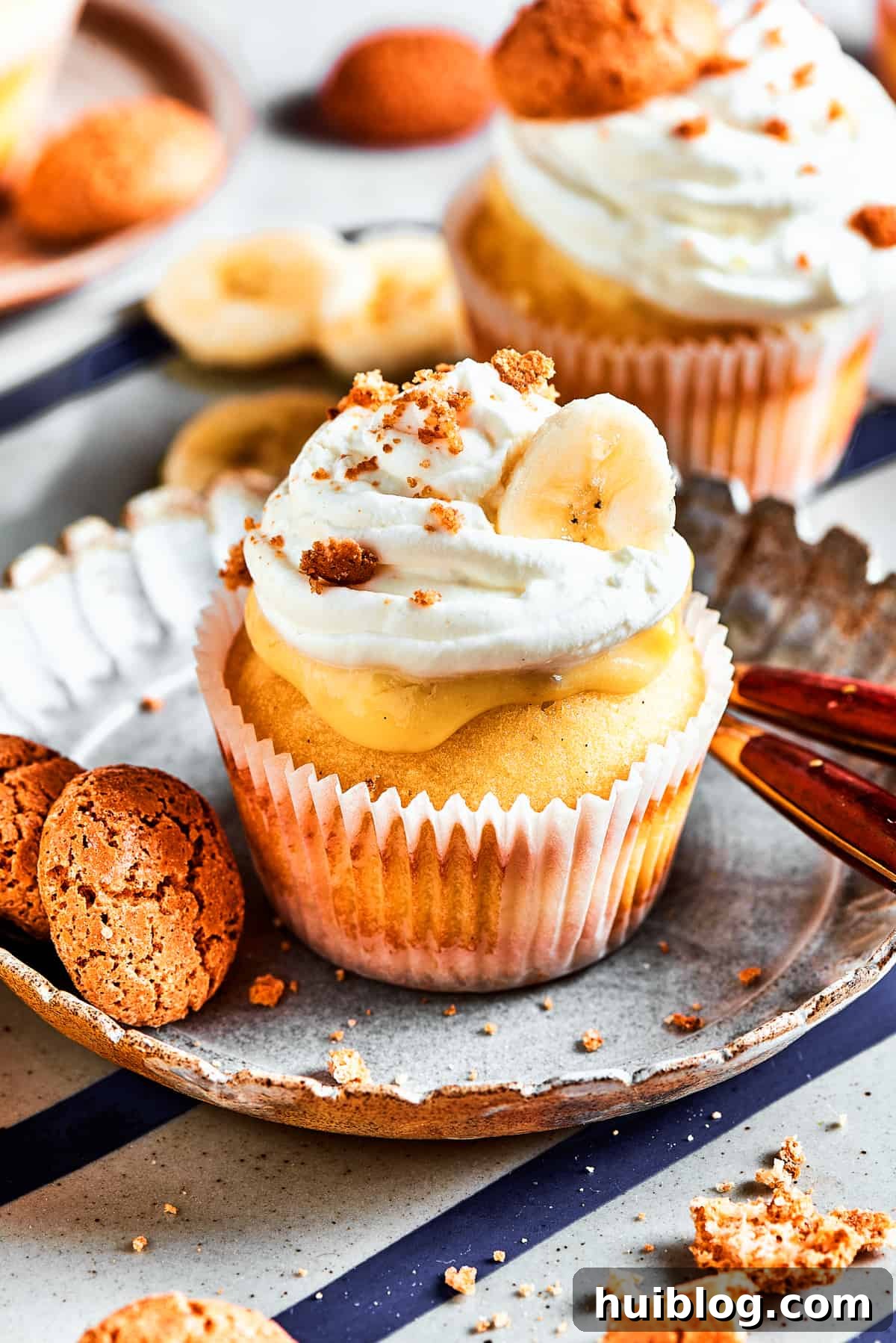Each Banana Pudding Cupcake is a delightful masterpiece, bringing joy with every bite! A single, beautifully adorned banana pudding cupcake rests on a dessert plate, with additional cupcakes subtly blurred in the background, highlighting its delightful details.