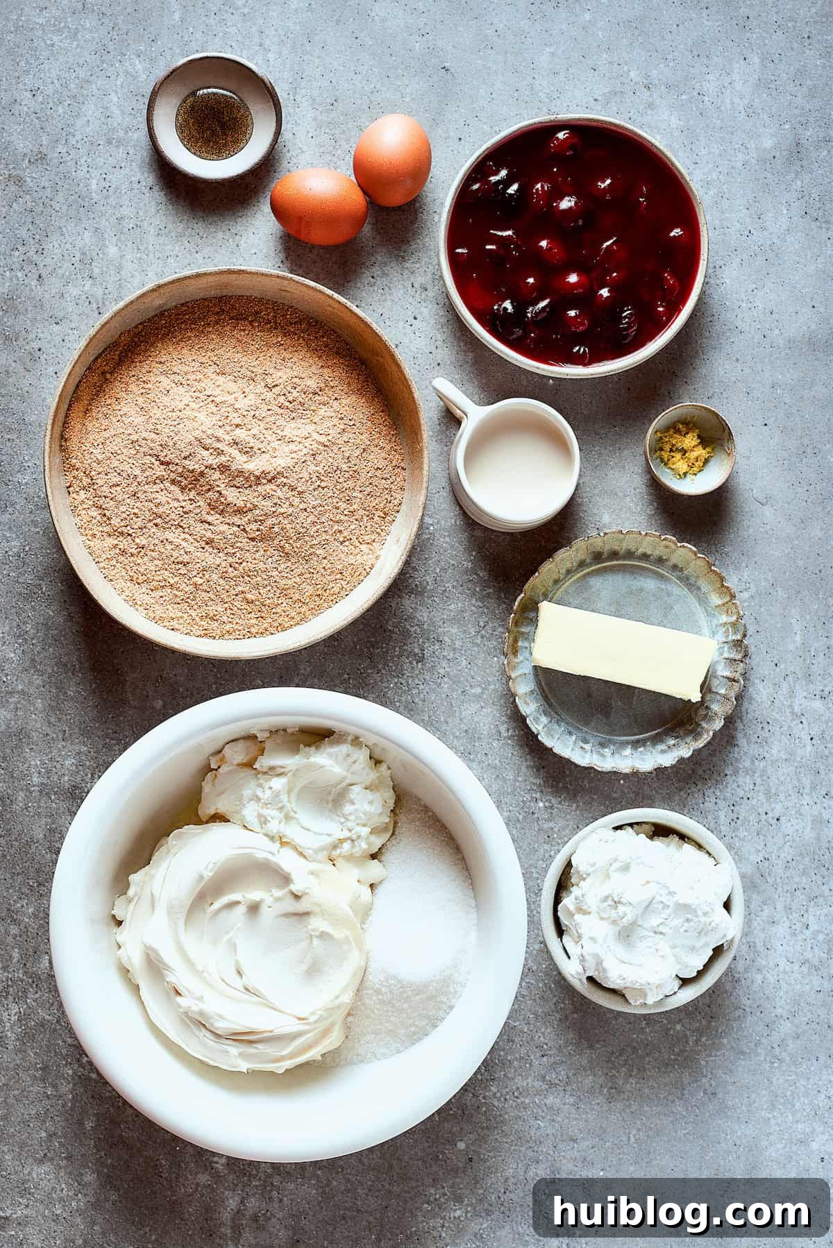 Fresh ingredients for making cherry cheesecake, including cream cheese, sour cream, eggs, graham crackers, butter, and cherry pie filling, artfully arranged on a rustic wooden surface.