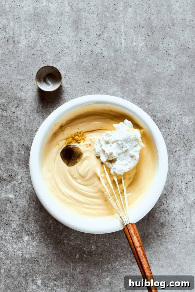 A person's hands operating a hand mixer to beat cream cheese and other ingredients together in a large mixing bowl for the cheesecake filling.