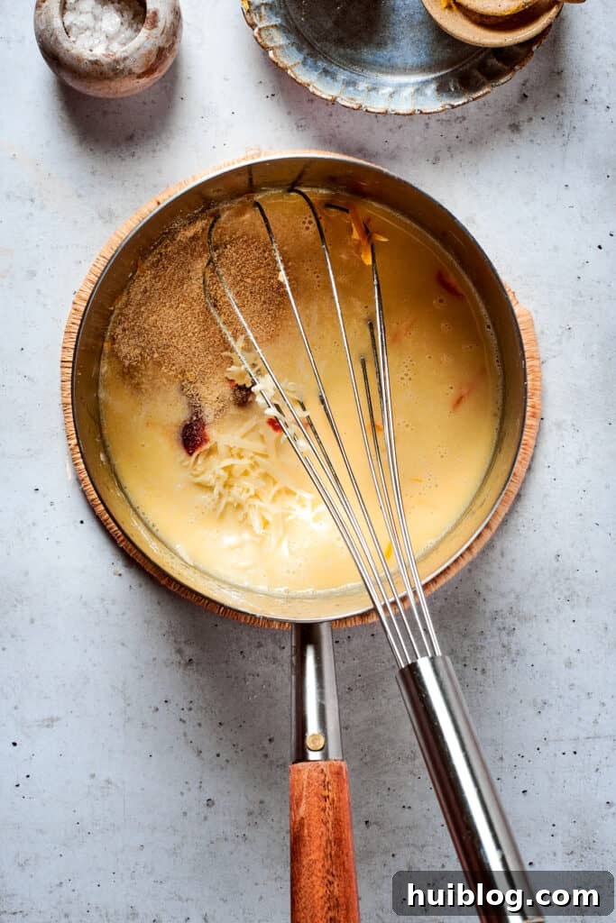 Close-up of a whisk blending a rich, velvety cheese sauce in a metal pot, highlighting the melted cheese and smooth consistency.