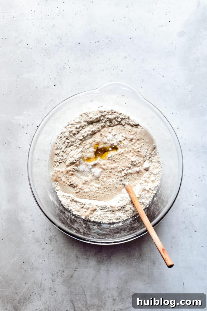 A glass bowl holds freshly mixed, slightly sticky soft pretzel dough, with a wooden spoon resting nearby.