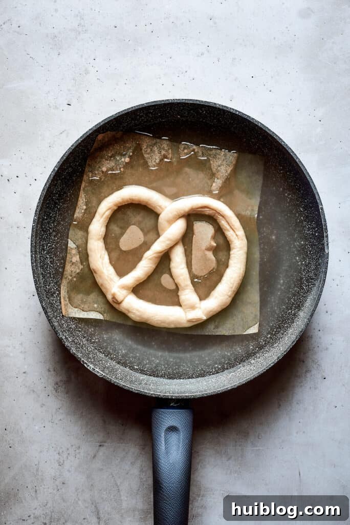 A raw, shaped soft pretzel is submerged in a pot of boiling water and baking soda, showing the crucial step of the baking soda bath.