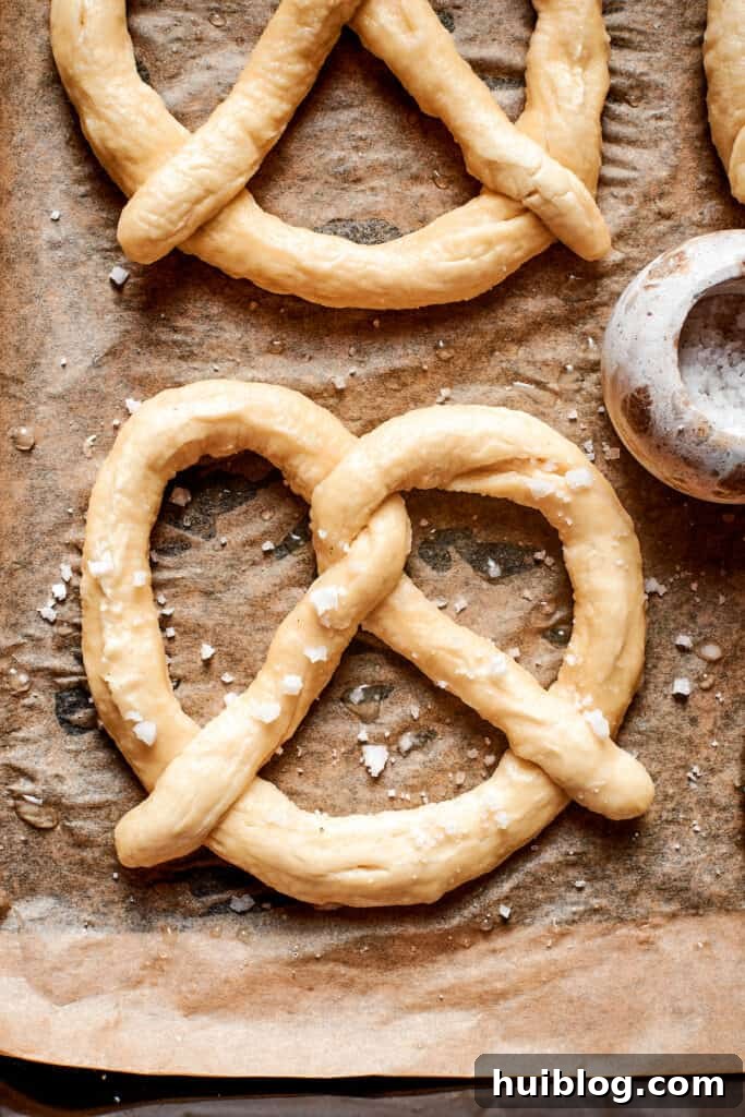 A baking sheet lined with parchment paper holds several raw, shaped soft pretzels, sprinkled with coarse salt, ready for the oven.